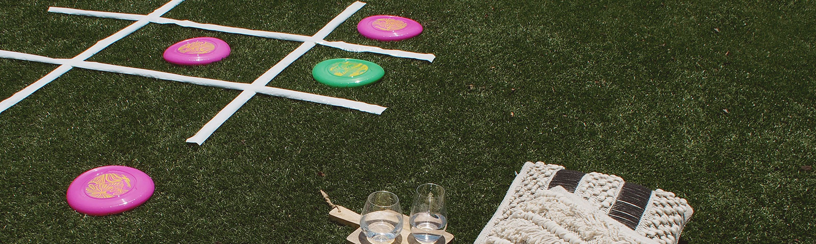 A large tic-tac-toe board sits on an artificial field. Frisbees are being used to mark the players and a blanket and pillows are setup nearby
