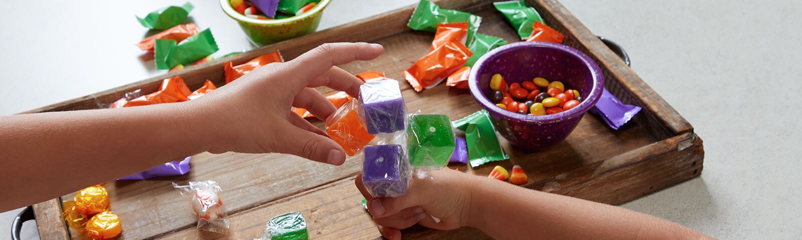 A rustic wooden tray covered in various candy in the background with a childs hand gripping lollipops and an adults hand reaching for them. A rustic wooden tray covered in various candy in the background with a childs hand gripping lollipops and an adults hand reaching for them.