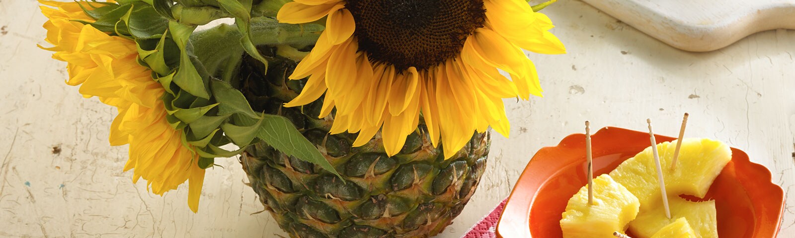 Close crop from above of a pineapple turned into a vase with large sunflowers sitting inside. To the right a plate with pineapple chunks.