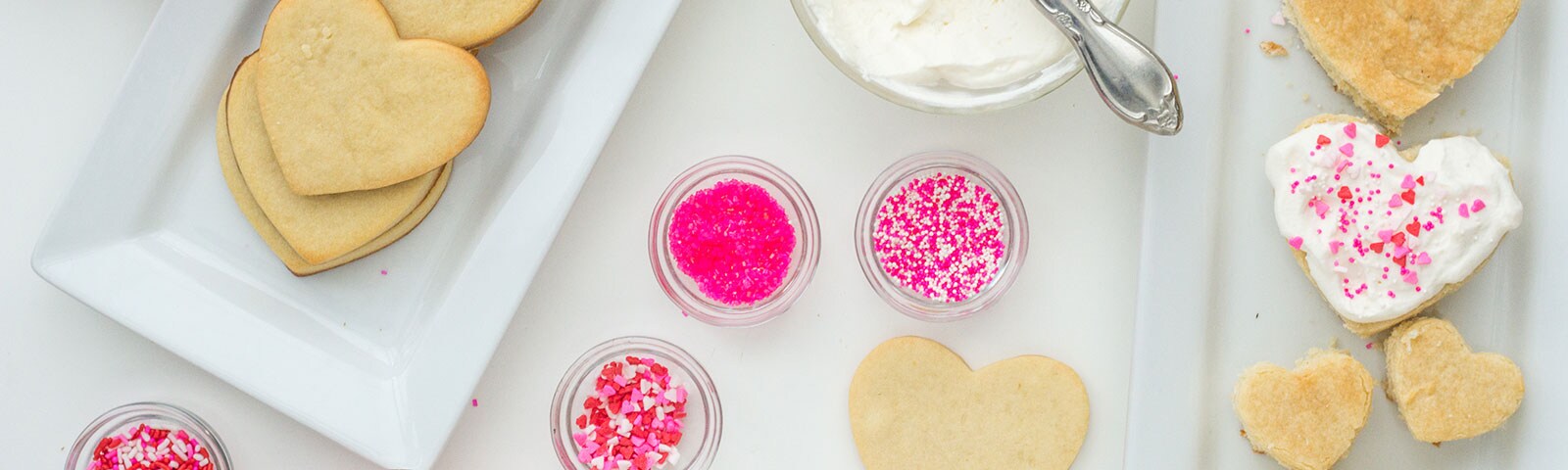Heart-shaped sugar cookies on a tray next to pink sprinkles and white frosting in separate bowls Heart-shaped sugar cookies on a tray next to pink sprinkles and white frosting in separate bowls